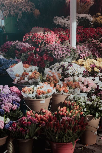 A vibrant outdoor display of various potted and arranged flowers at a florist market stands. The assortment includes pink, white, and purple chrysanthemums, pink tulips, and peach-colored roses, all a