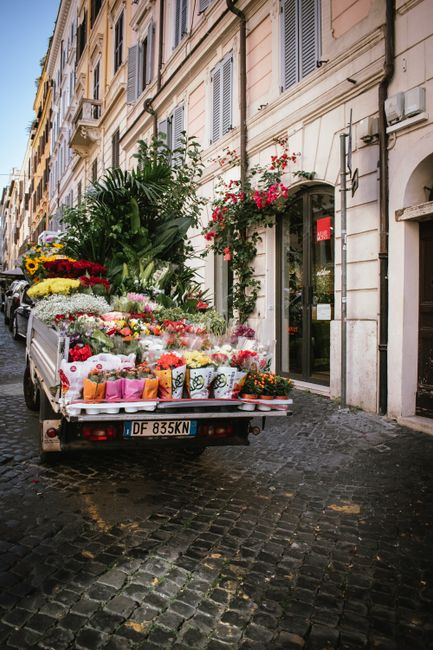 A delivery truck parked on a cobblestone street in front of a classic European building with cream-colored walls and green window shutters. The truck's cargo bed is filled with an assortment of fresh 